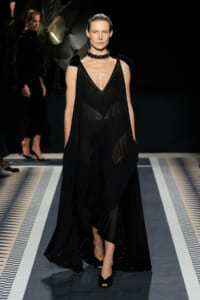 Model walks the runway in a black layered gown with a deep V, sheer panels, and cape-like shoulders; sleek choker necklace completes the look.