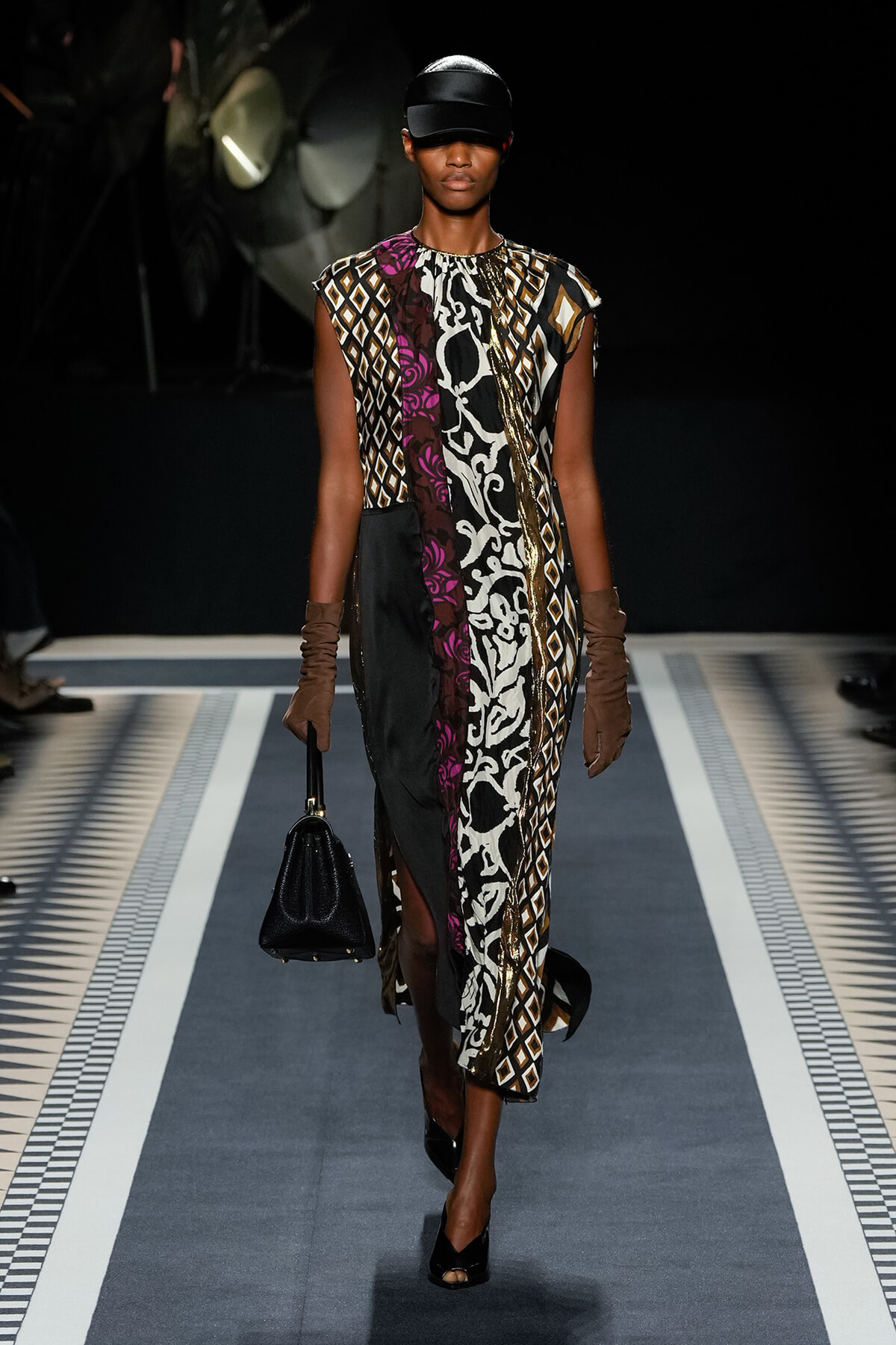 Model walking a fashion runway in a mixed-pattern sleeveless dress, black cap, brown gloves, and a black handbag.