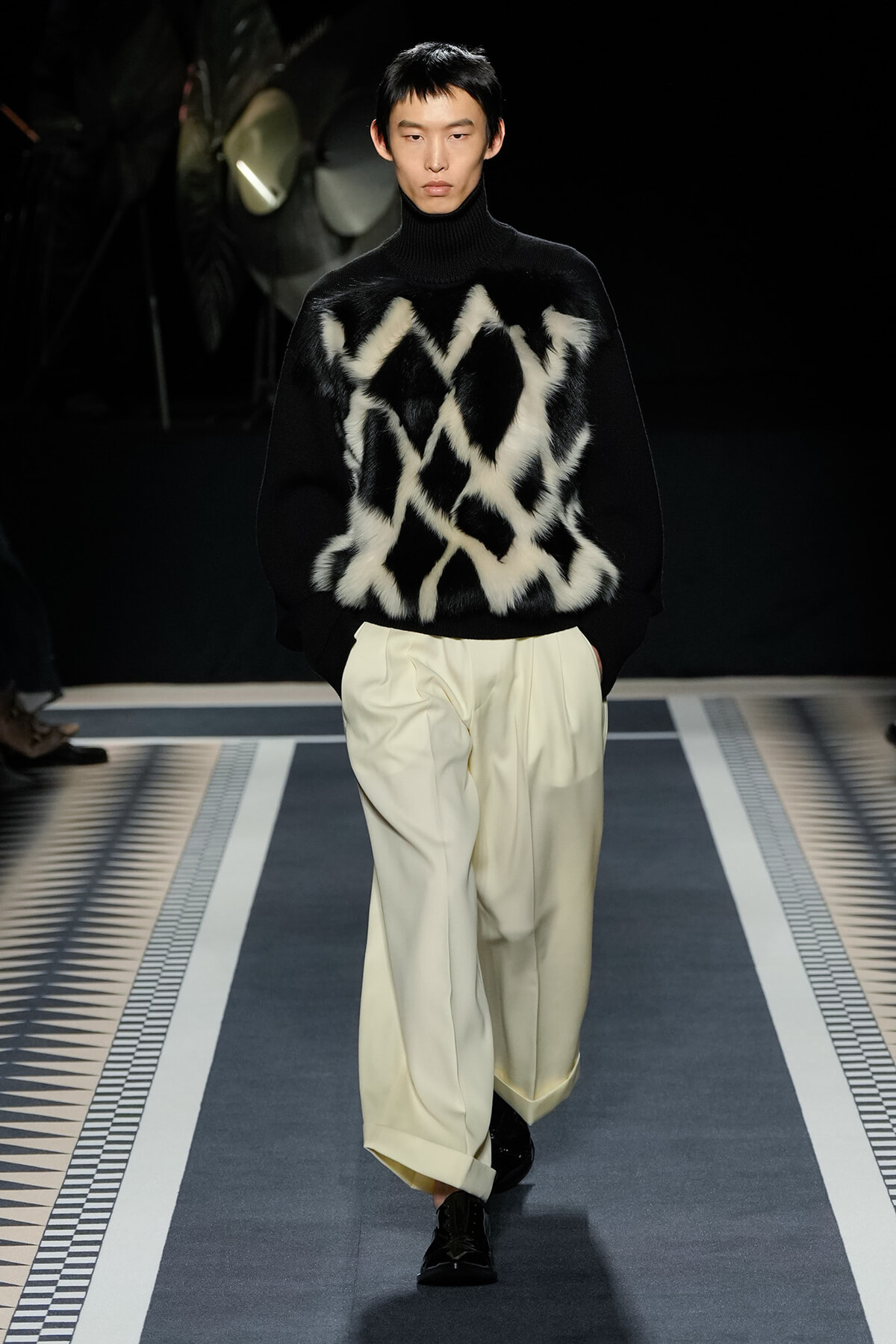 Runway model in a black turtleneck with white fur geometric panels and cream wide-leg trousers, black shoes.