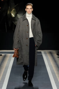 Male fashion model on a runway wearing a oversized checked overcoat, white turtleneck, black trousers, and black loafers, carrying a brown clutch bag