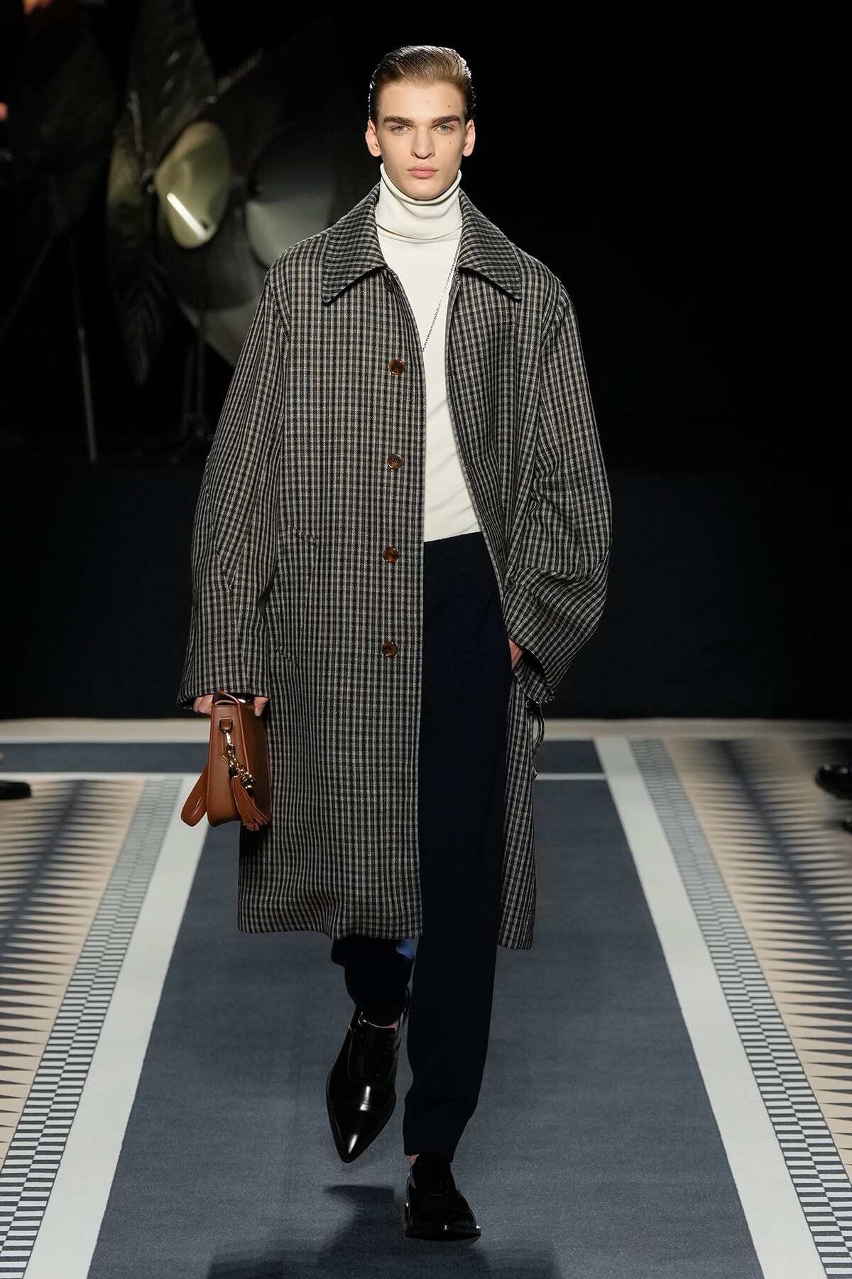 Male fashion model on a runway wearing a oversized checked overcoat, white turtleneck, black trousers, and black loafers, carrying a brown clutch bag