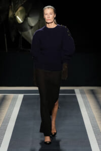 Model walking a fashion runway in a navy knit sweater with voluminous sleeves, black skirt, and brown gloves against a dark backdrop.