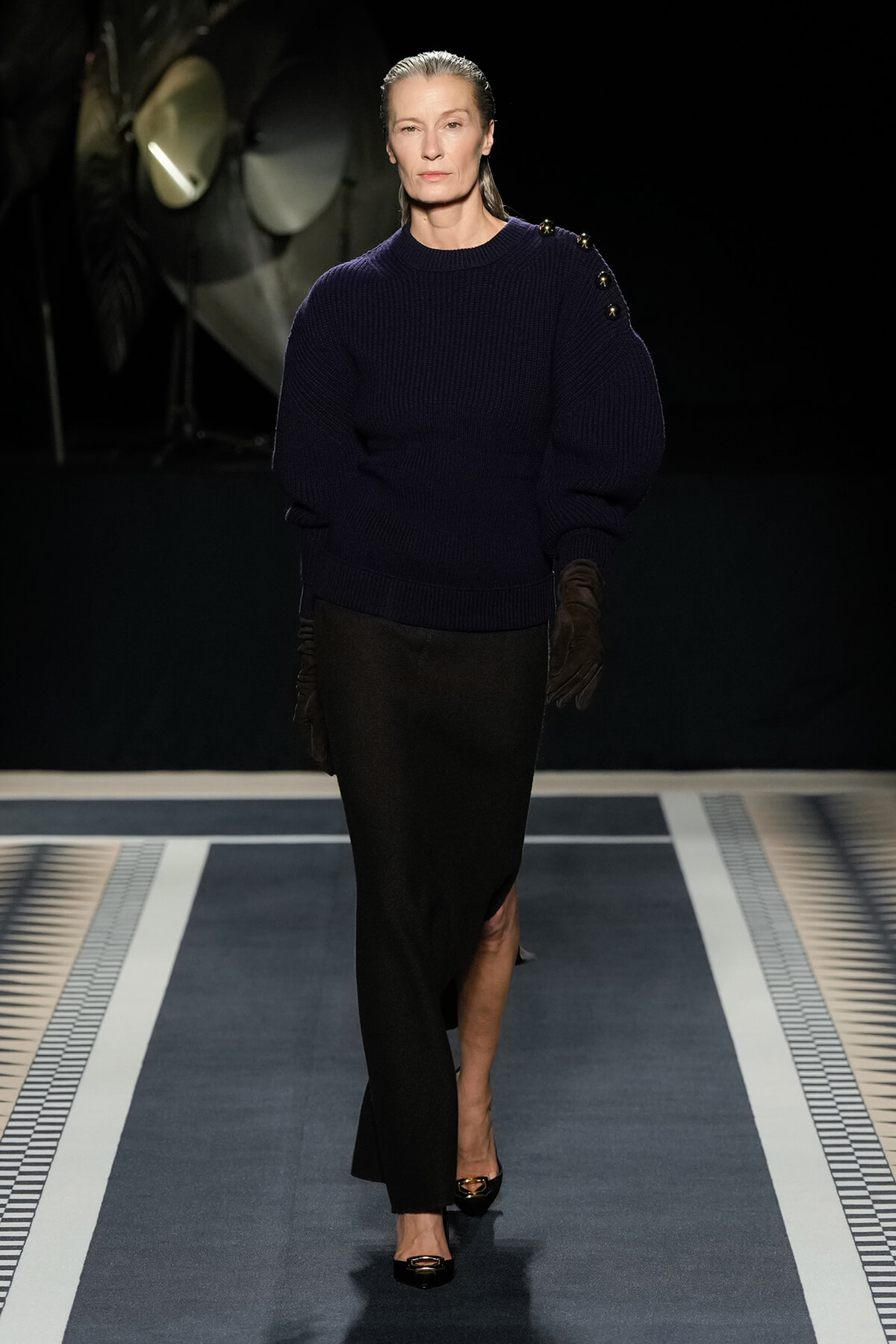 Model walking a fashion runway in a navy knit sweater with voluminous sleeves, black skirt, and brown gloves against a dark backdrop.