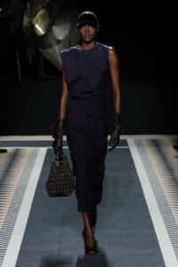 Model walks the runway in a navy sleeveless wrap dress, black gloves, and a dark cap, carrying a perforated black handbag.