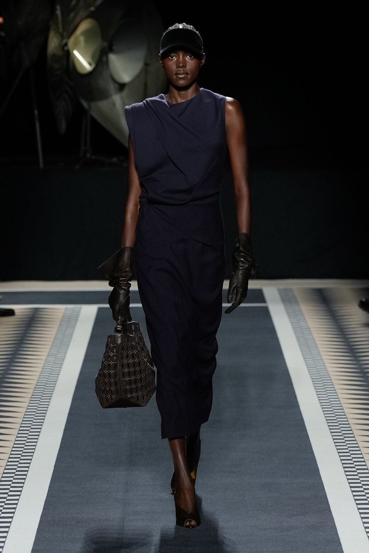Model walks the runway in a navy sleeveless wrap dress, black gloves, and a dark cap, carrying a perforated black handbag.