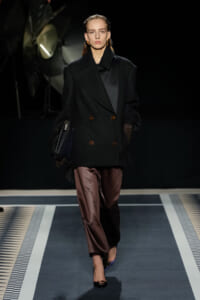 Model walking a runway in an oversized black double-breasted coat, brown satin trousers, and black top, carrying a black bag and wearing brown gloves.