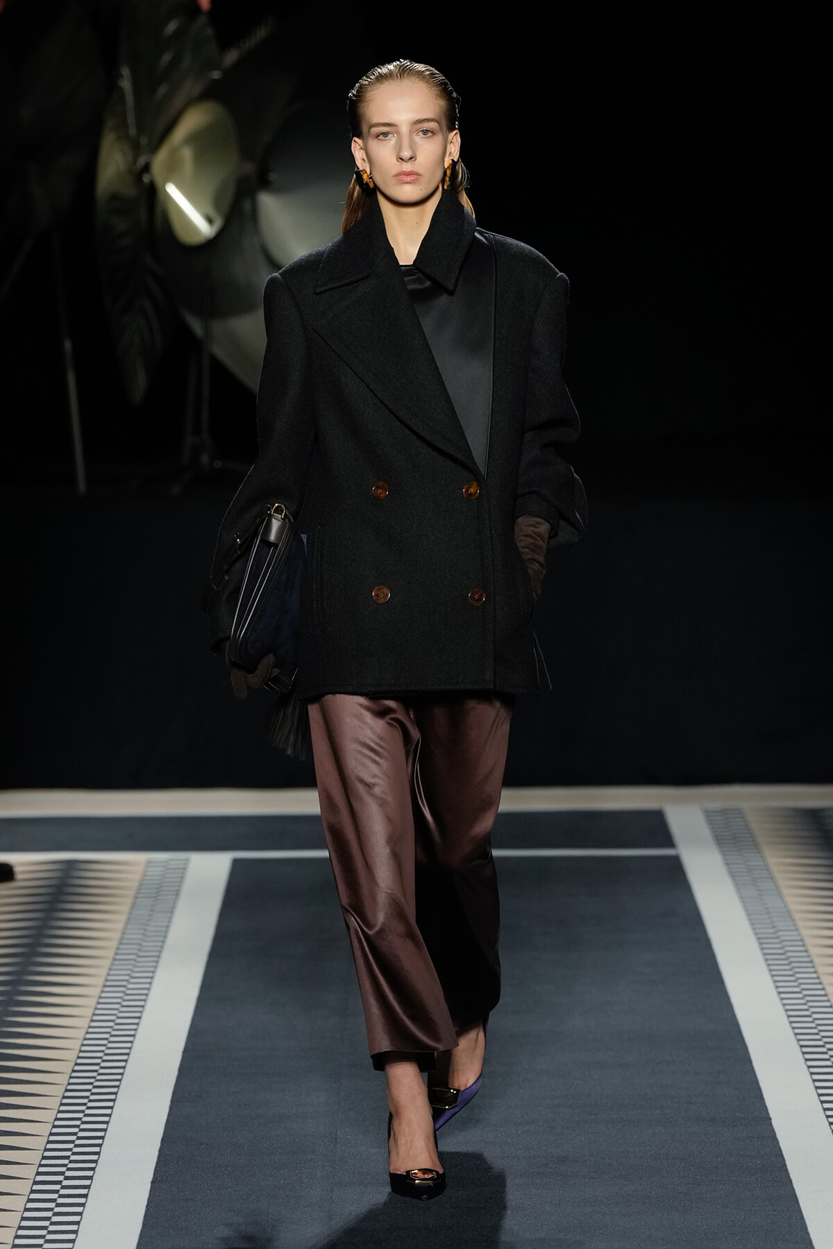 Model walking a runway in an oversized black double-breasted coat, brown satin trousers, and black top, carrying a black bag and wearing brown gloves.