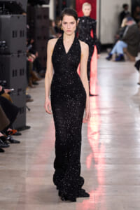 Runway model walks in a floor-length black sequined halter gown with a subtle train, during a fashion show.