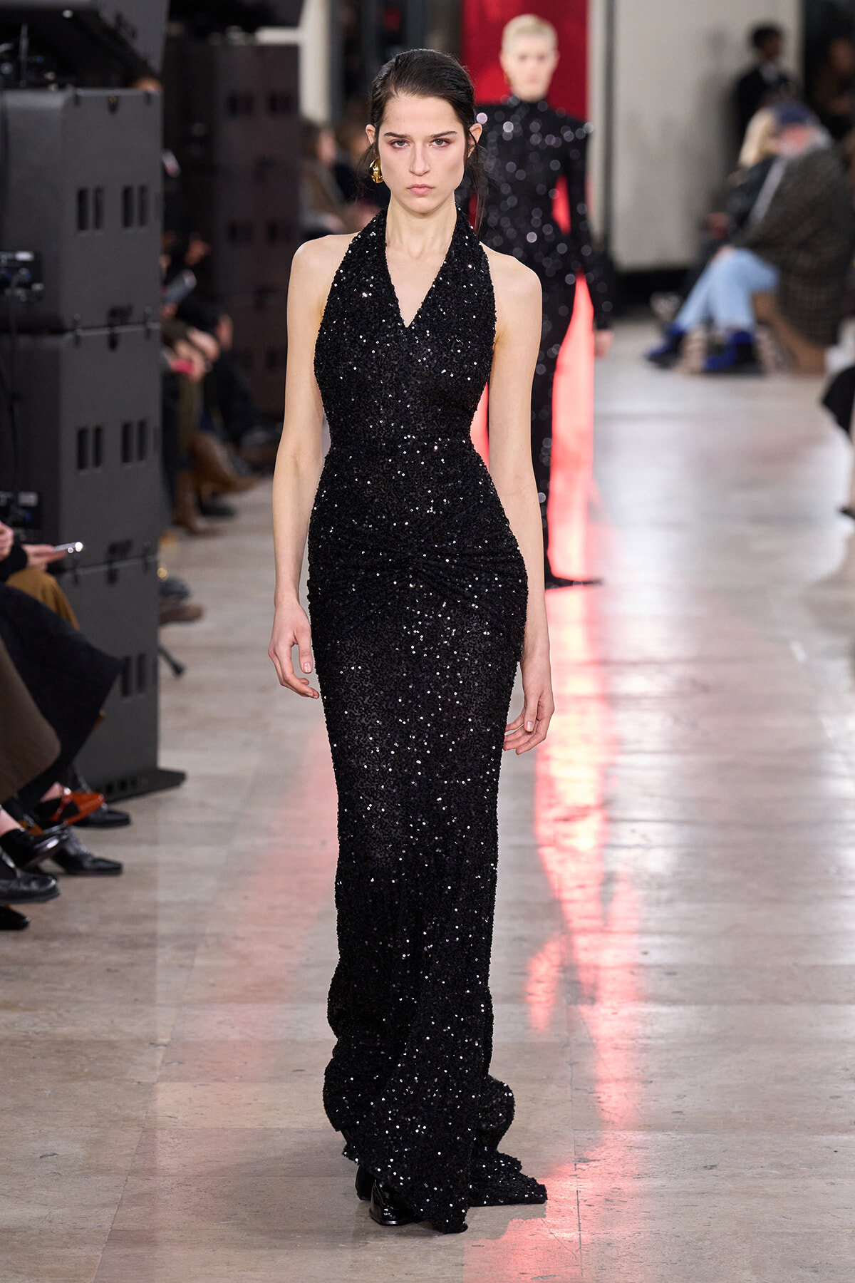 Runway model walks in a floor-length black sequined halter gown with a subtle train, during a fashion show.