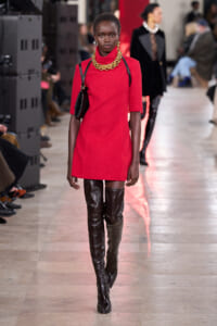 Model walking a fashion runway in a bright red mini dress, oversized gold chain necklace, and thigh-high glossy black boots with a black shoulder bag.