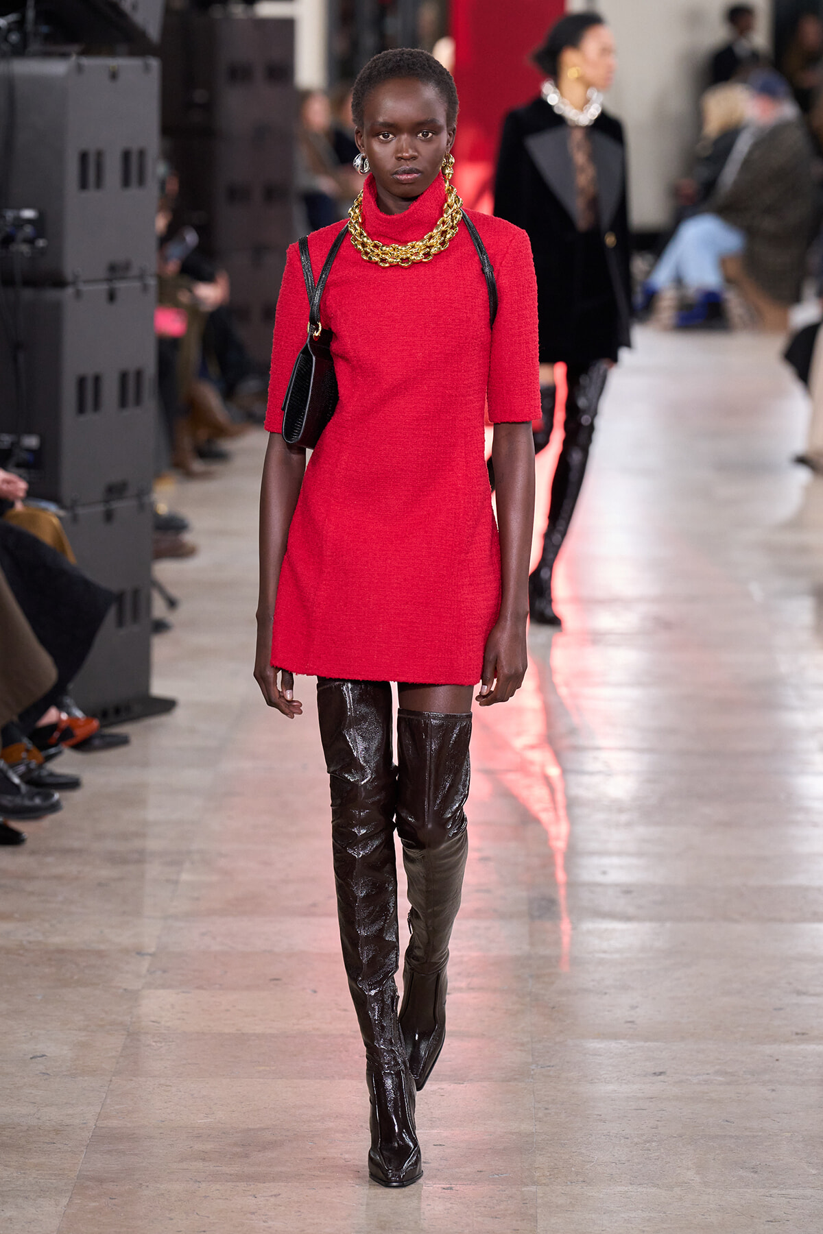 Model walking a fashion runway in a bright red mini dress, oversized gold chain necklace, and thigh-high glossy black boots with a black shoulder bag.