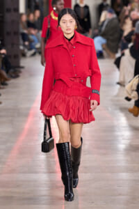 Runway model in a red textured coat with a wide belt, matching red skirt, black knee-high boots, and a small black handbag on a fashion show runway.
