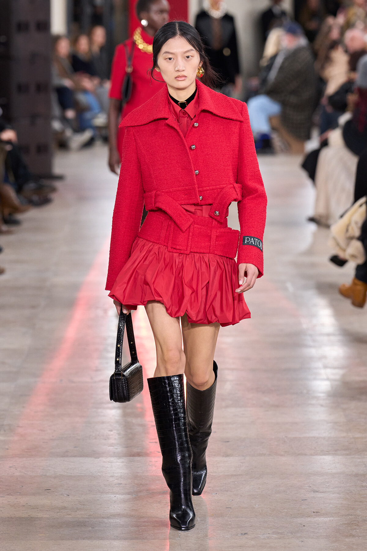 Runway model in a red textured coat with a wide belt, matching red skirt, black knee-high boots, and a small black handbag on a fashion show runway.