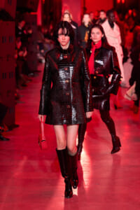 Model walks the runway in a shiny black crocodile-patterned coat with knee-high boots under red lighting, holding a red bag.