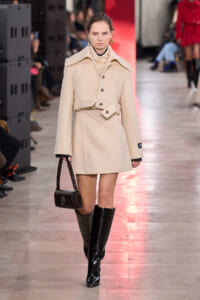 Model walking a fashion runway in a cream textured coat with wide collar, belt, and black knee-high boots, carrying a small dark handbag.