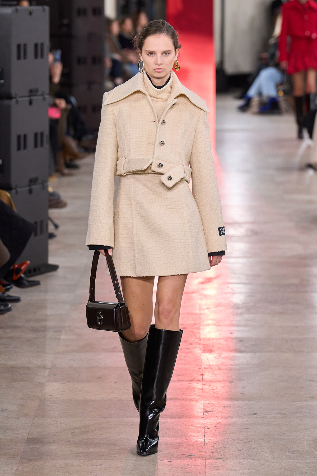 Model walking a fashion runway in a cream textured coat with wide collar, belt, and black knee-high boots, carrying a small dark handbag.