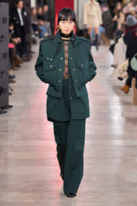 Model on a runway wearing a dark green utility jacket with gold buttons and matching wide-leg pants; lace top visible at the neckline.