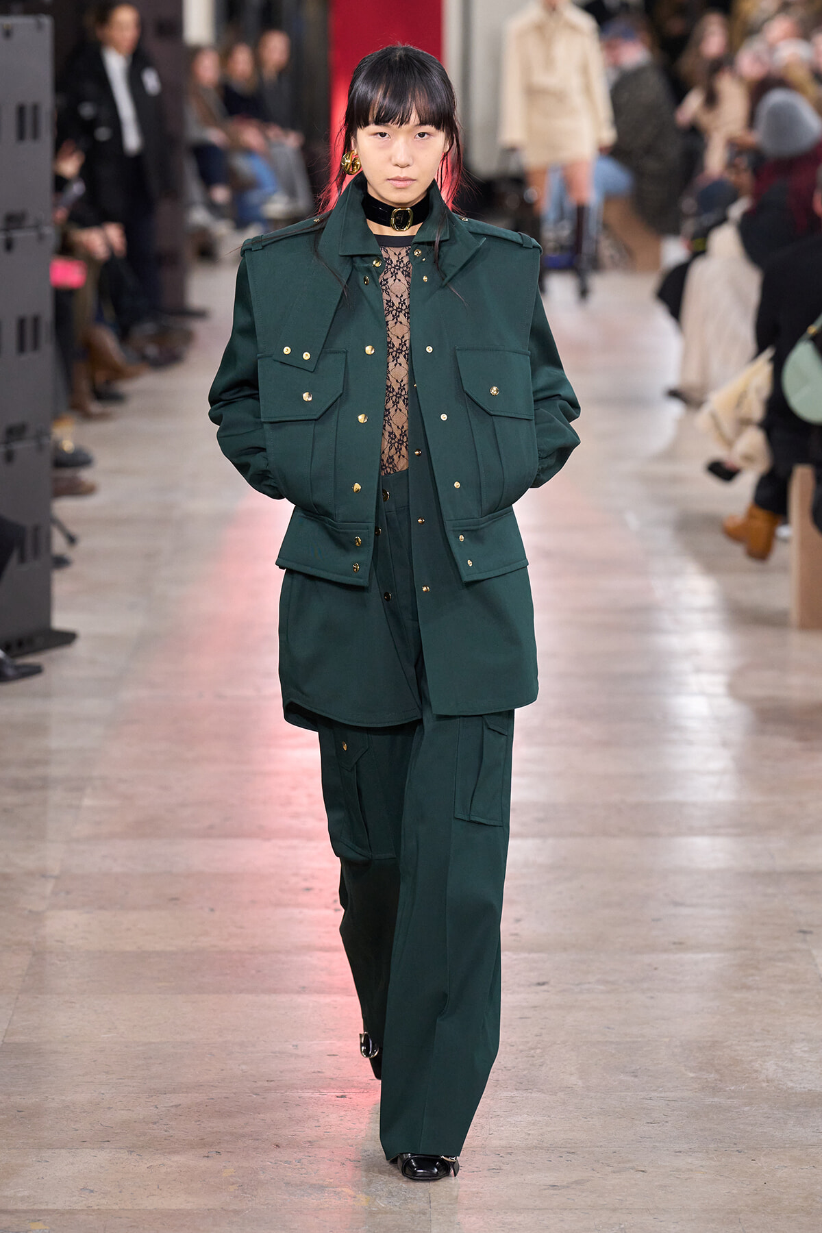 Model on a runway wearing a dark green utility jacket with gold buttons and matching wide-leg pants; lace top visible at the neckline.