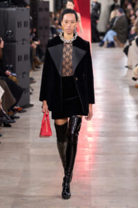 Model walking a runway in a black velvet blazer over a sheer lace top, black mini skirt, and thigh-high patent boots, carrying a red bag.