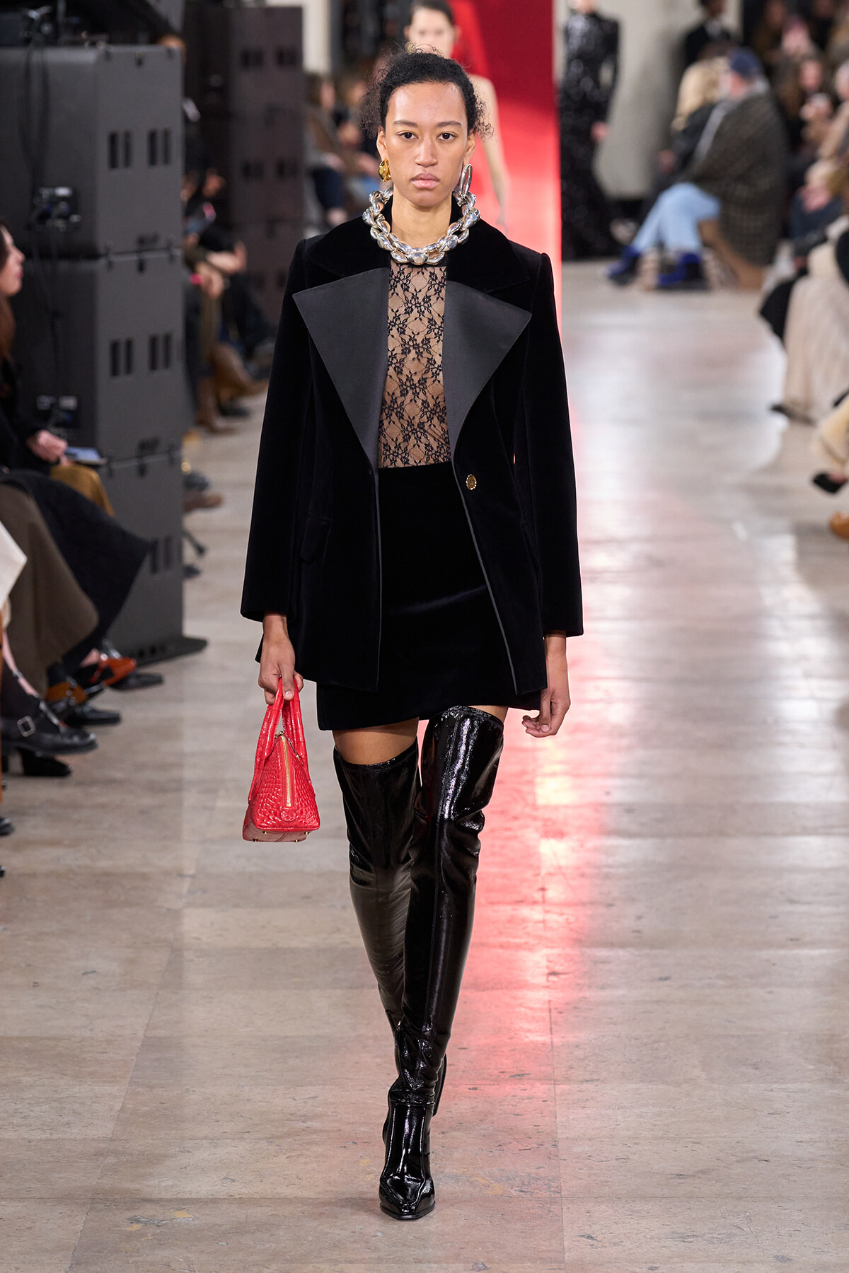 Model walking a runway in a black velvet blazer over a sheer lace top, black mini skirt, and thigh-high patent boots, carrying a red bag.