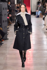 Female model on a runway wearing a black trench coat with a white oversized collar, beige striped neck scarf, wide black crocodile-embossed belt, and knee-high patent boots.