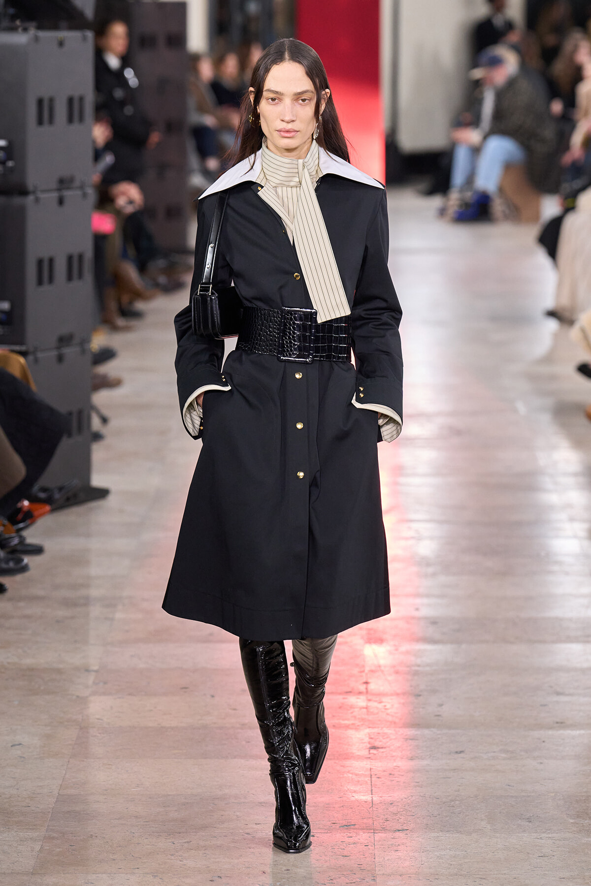 Female model on a runway wearing a black trench coat with a white oversized collar, beige striped neck scarf, wide black crocodile-embossed belt, and knee-high patent boots.