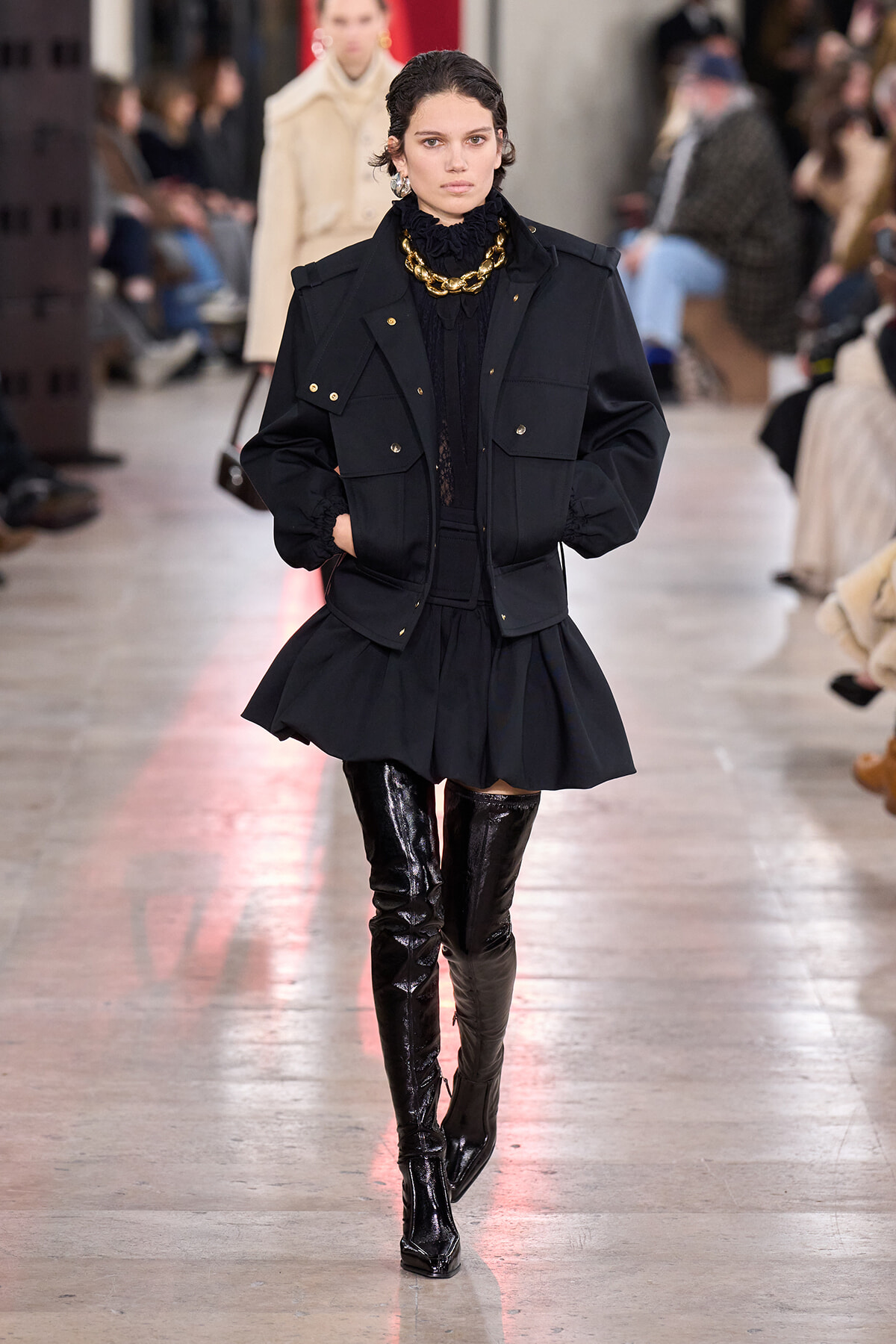 Model on a fashion runway wearing a black utility-style jacket, black ruffled top, short flared skirt and glossy black over-the-knee boots with a chunky gold necklace.