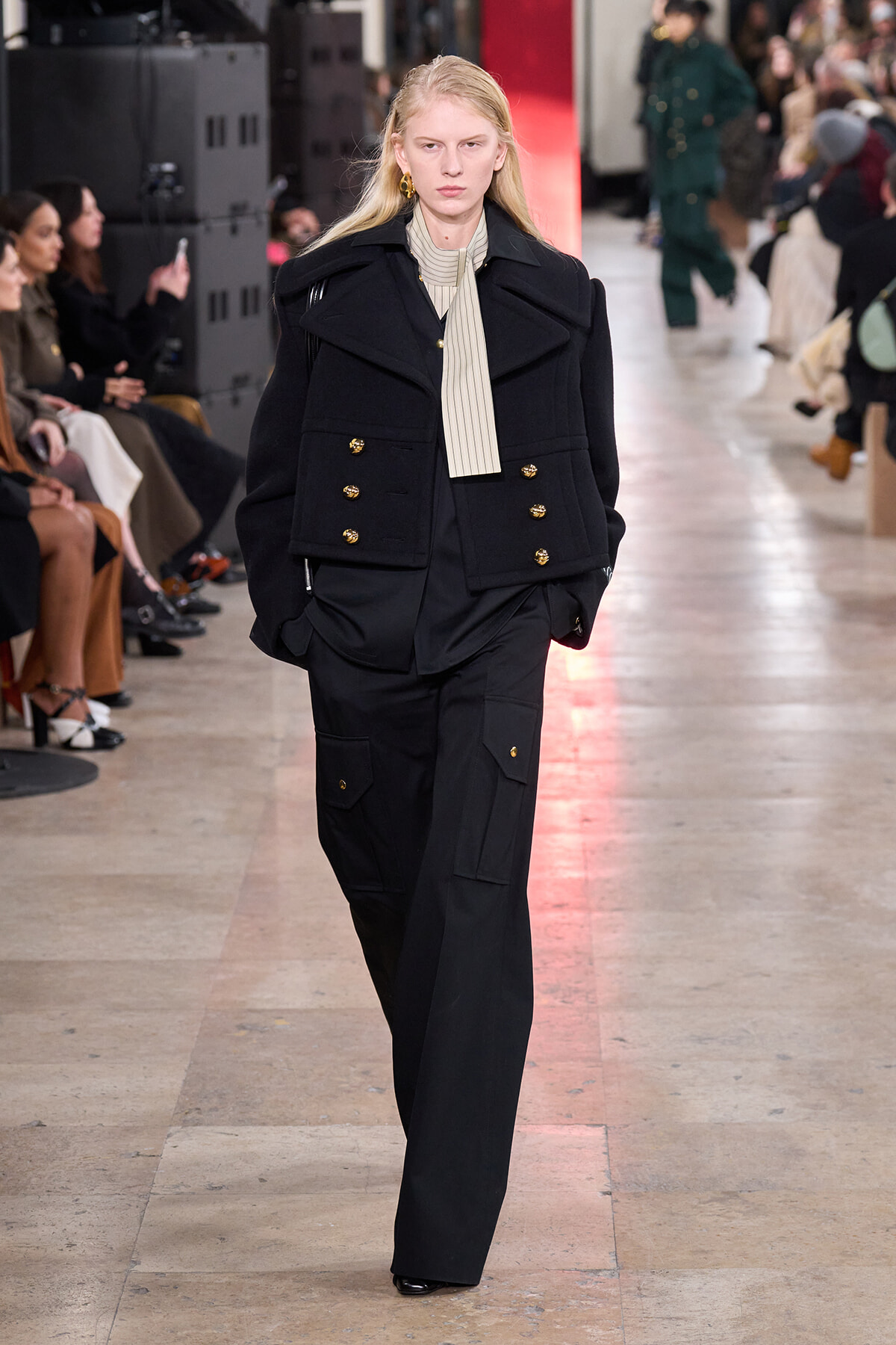 Model walking a runway in a black double-breasted jacket with gold buttons, beige striped scarf, and black cargo pants.