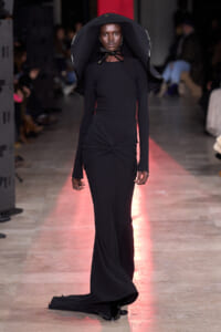 Model walks the runway in a form-fitting black gown with a twist at the waist and a large, sculptural black hat shaped like a brimmed cape.