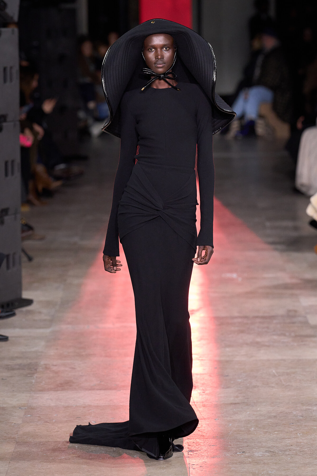 Model walks the runway in a form-fitting black gown with a twist at the waist and a large, sculptural black hat shaped like a brimmed cape.