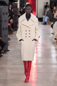 Model on a runway wearing a cream double-breasted wool coat with wide lapels and red knee-high boots.
