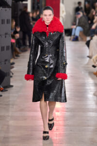 Model walking a runway in a black crocodile-embossed trench with a red fluffy collar and cuffs, belted at the waist.