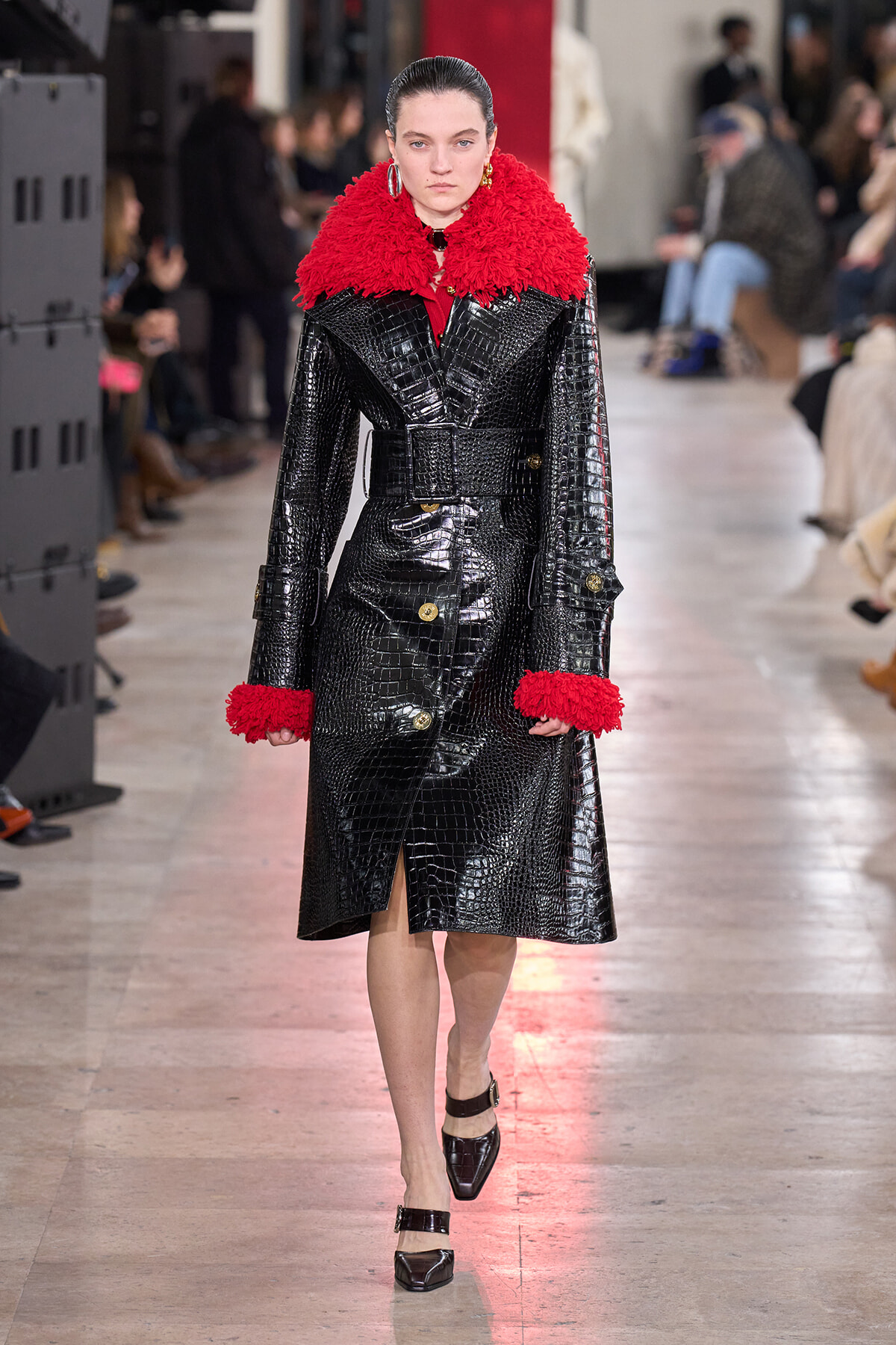 Model walking a runway in a black crocodile-embossed trench with a red fluffy collar and cuffs, belted at the waist.