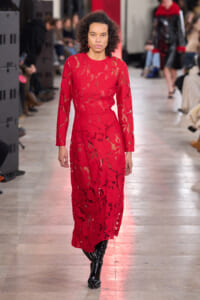 Model walking the runway in a full-length red lace dress with long sleeves and black boots, on a fashion show catwalk with a focused expression.