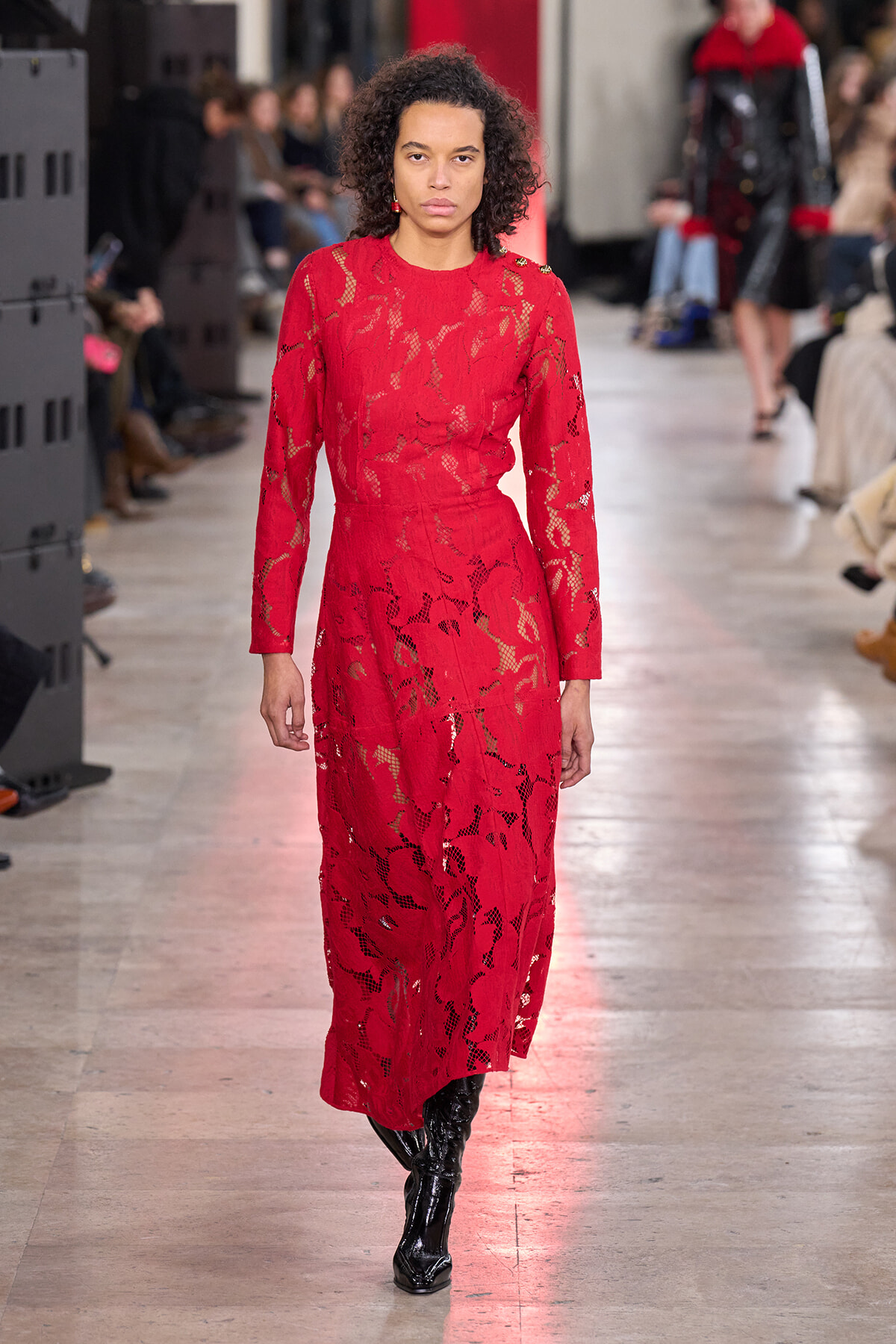 Model walking the runway in a full-length red lace dress with long sleeves and black boots, on a fashion show catwalk with a focused expression.