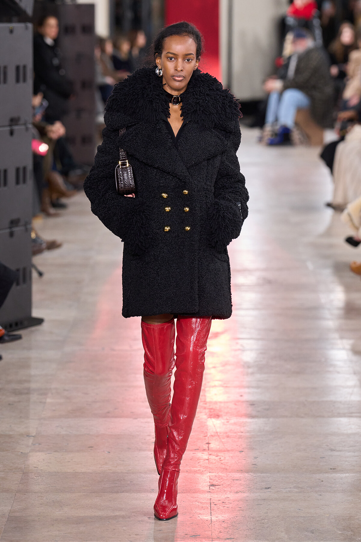 Model walks the runway in a black fluffy, double-breasted coat with gold buttons and red knee-high boots.