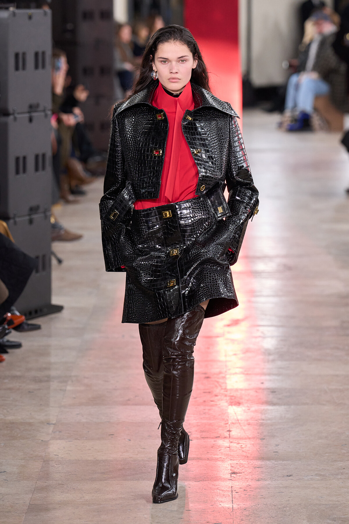 Model walks the runway in a shiny black crocodile-embossed coat over a red blouse, with matching knee-high boots in a fashion show setting.