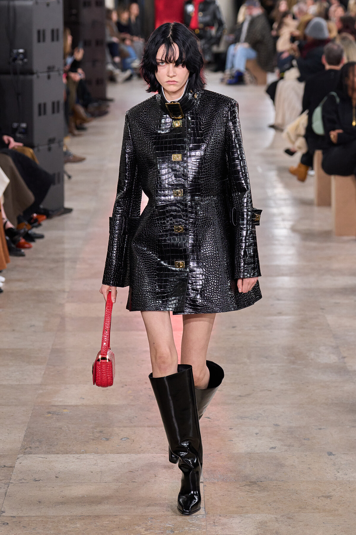 Model walking a runway in a glossy black crocodile-embossed coat with gold buckles, paired with knee-high black boots and a small red handbag.