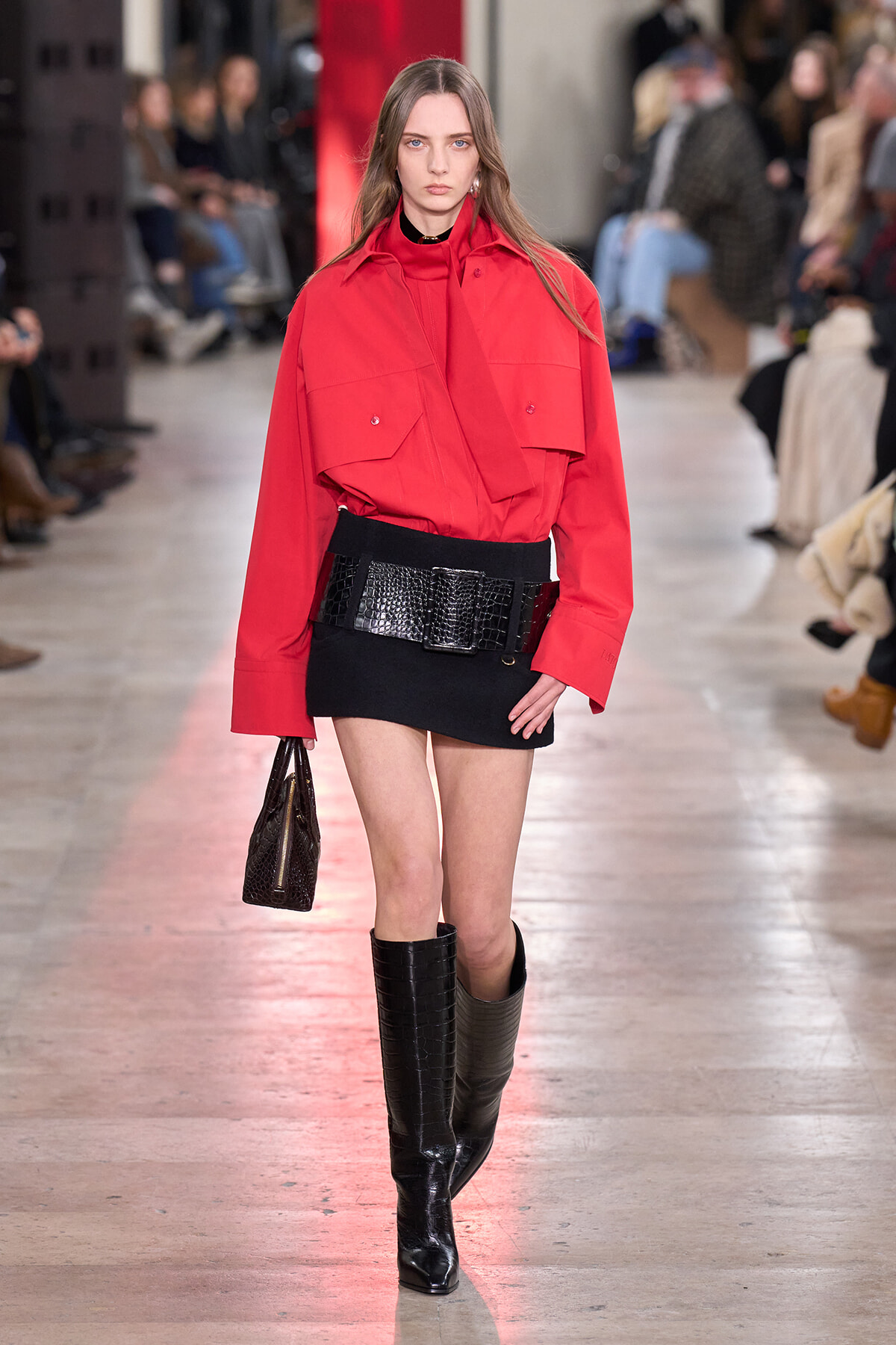 Model on runway wearing a bright red oversized jacket, black mini skirt, wide black belt, and tall black boots while carrying a textured clutch.