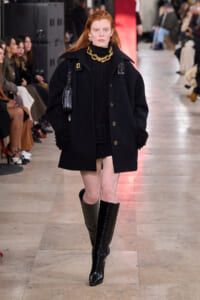 Model on runway wearing an oversized black coat with gold buttons, a black turtleneck, a chunky gold chain, and knee-high black boots.
