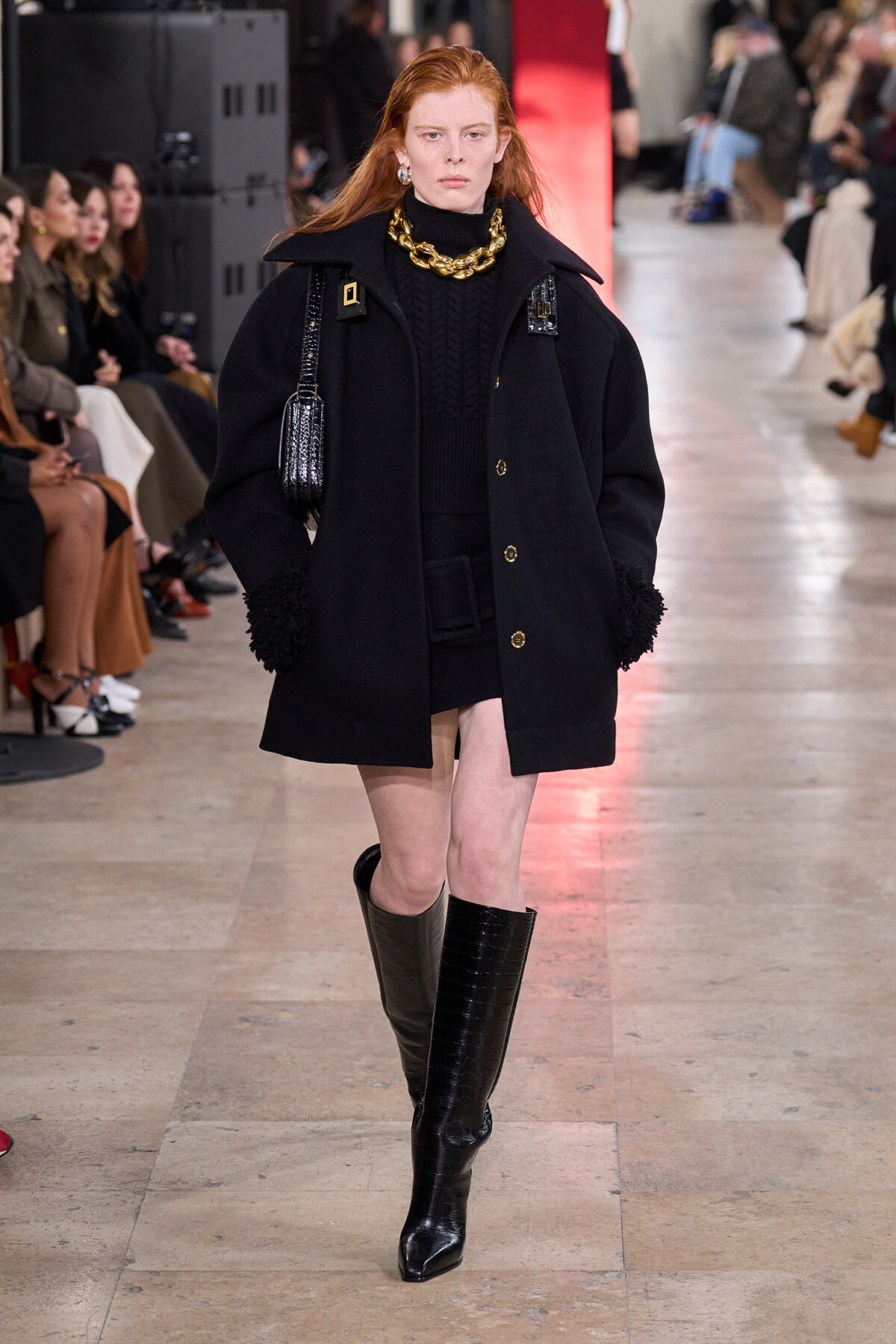 Model on runway wearing an oversized black coat with gold buttons, a black turtleneck, a chunky gold chain, and knee-high black boots.