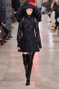 Runway model in a black lace dress with an oversized curved hat and glossy thigh-high boots.