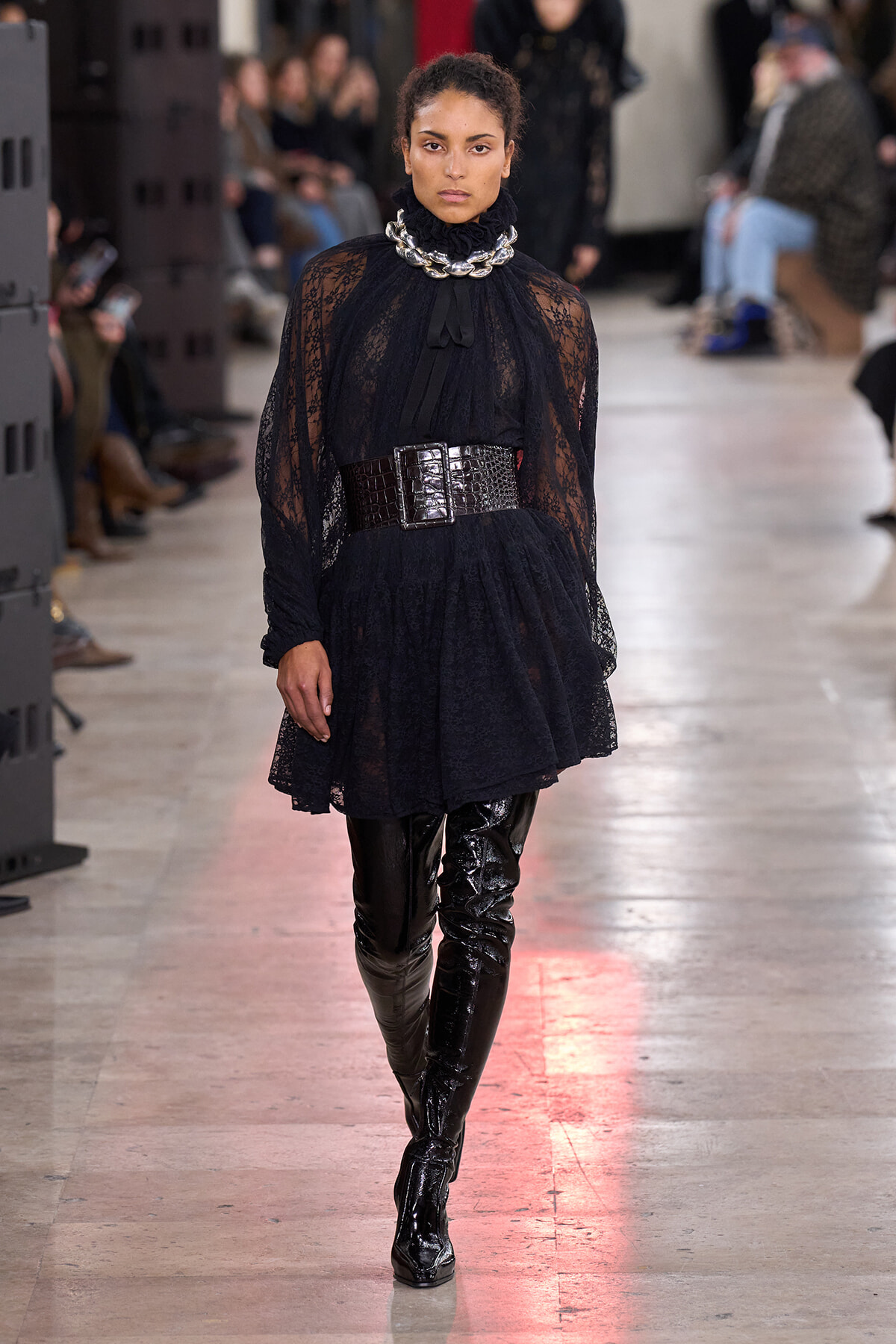 Model walking runway in black lace tunic dress with oversized metallic belt and glossy thigh-high boots; fashion show setting.