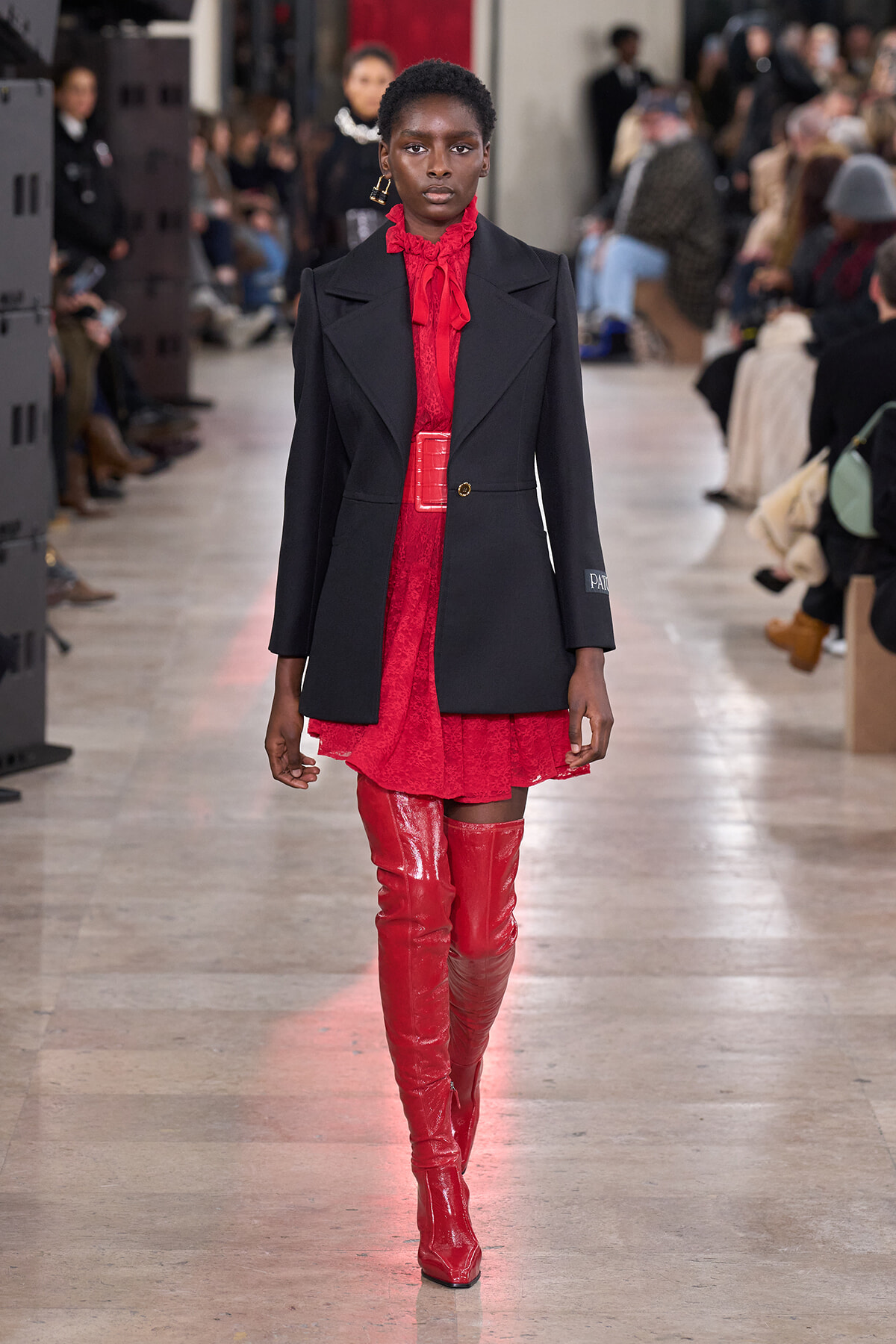 Model walks the runway in a red lace dress with a black blazer, red belt, and matching red knee-high boots.