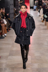 Model walks a fashion runway in an oversized black coat with vertical branding, paired with a red ruffled blouse and glossy black knee-high boots.