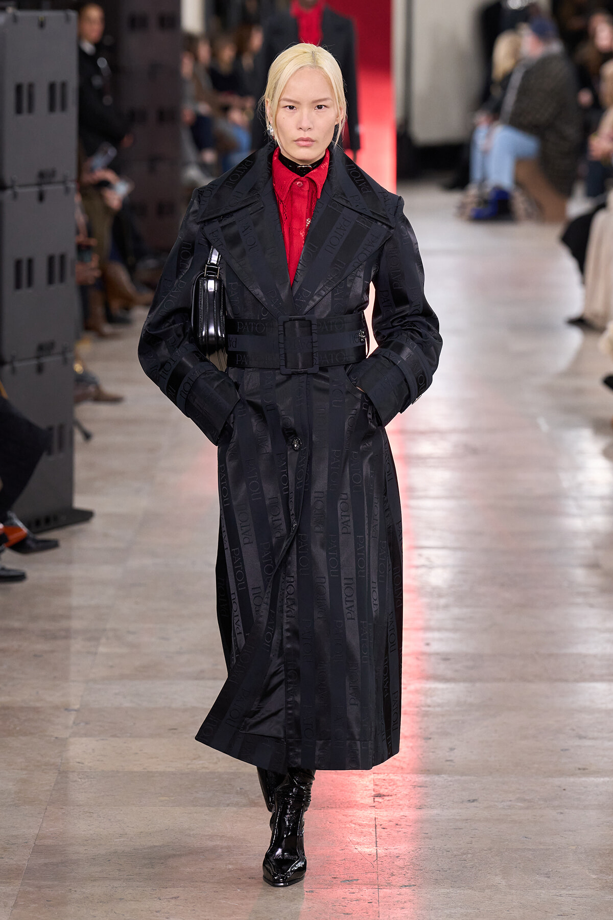 Model walking a fashion show in a long black trench coat with a wide belt over a red blouse, black patent boots, and a small black bag over the shoulder; audience blurred in the background.