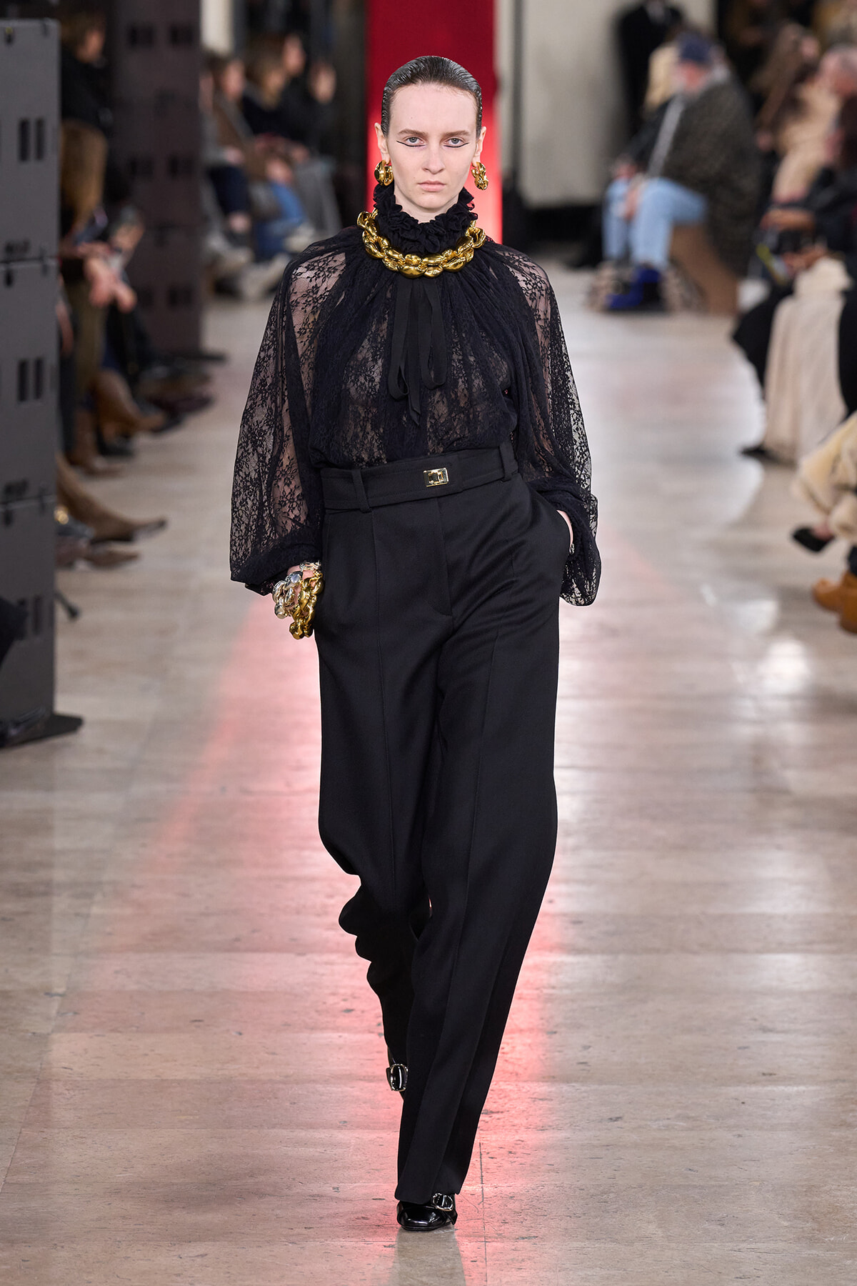 Model walking a runway in a sheer black lace blouse with a high neck and chunky gold jewelry, paired with tailored black trousers.