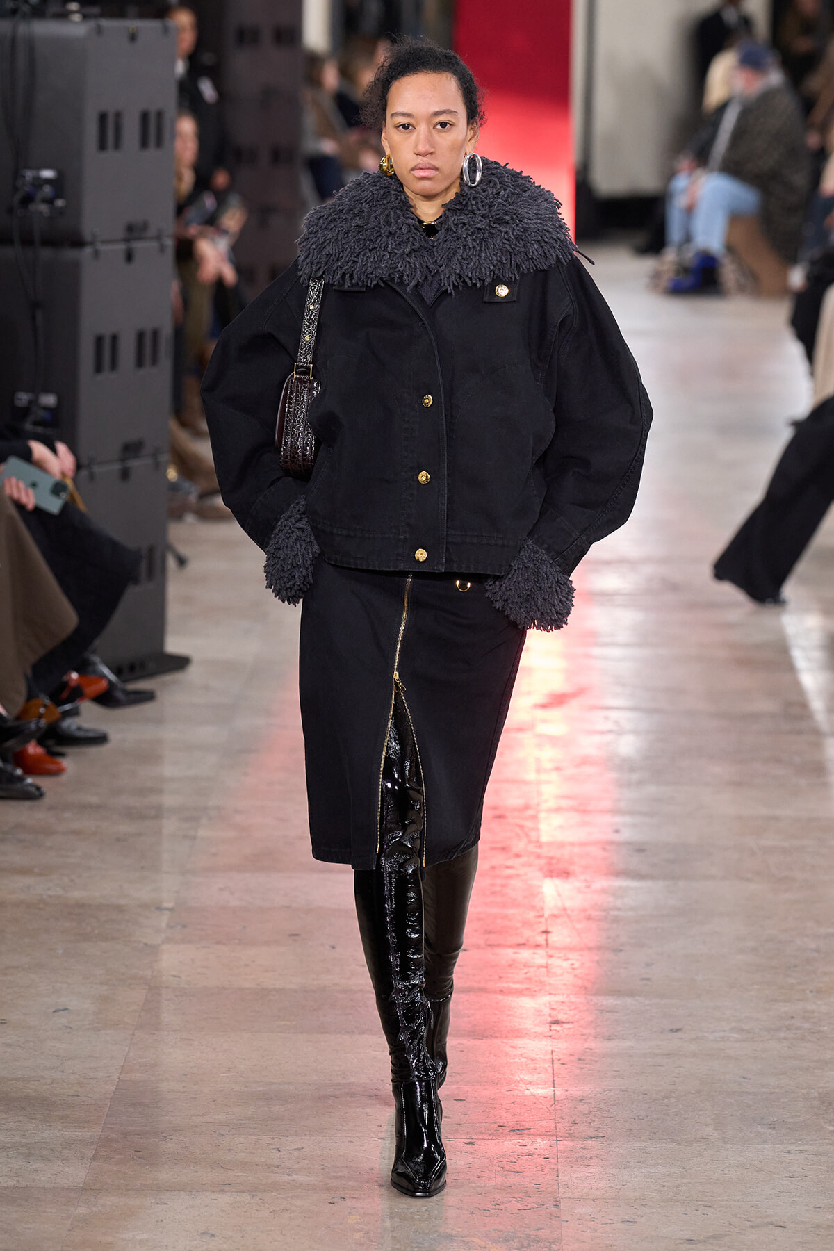 Model on a runway wearing an oversized black jacket with a fluffy shearling collar and cuffs, gold buttons, a black midi skirt with a front zipper, and tall black patent boots, carrying a small shoulder bag.