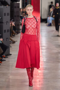 Model walking a runway in a sheer red lace top, red high-waisted skirt with a wide belt, and red knee-high boots.