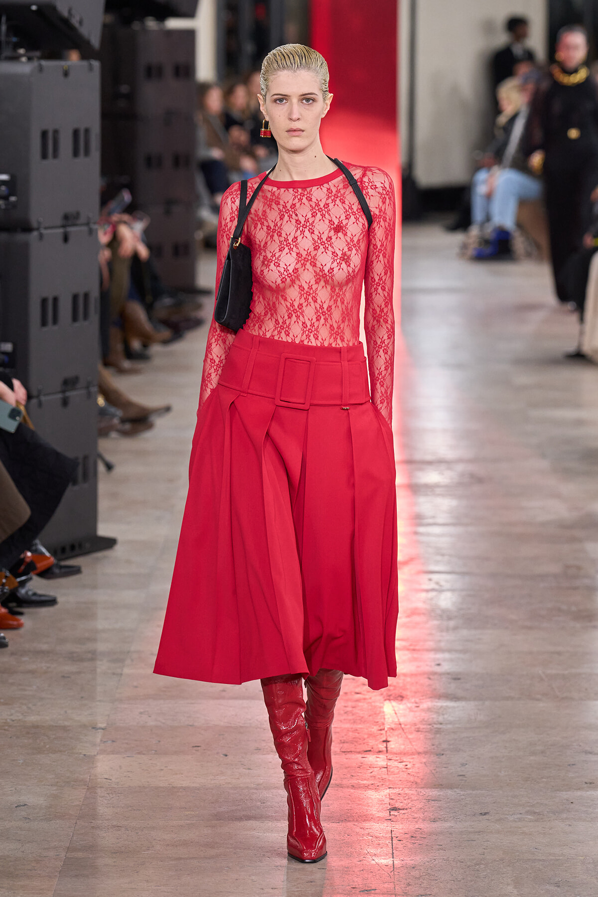 Model walking a runway in a sheer red lace top, red high-waisted skirt with a wide belt, and red knee-high boots.